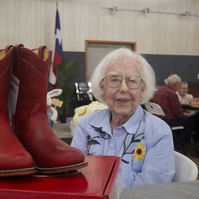 A lady with gray hair, glasses, and a blue and white striped shirt with flowers posing and smiling next to her first pair of red cowboy roper boots. 